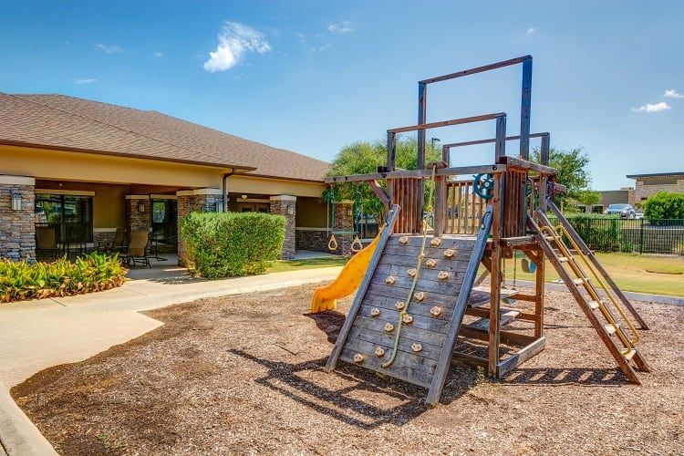 A sunny playground with a climbing structure and wood chips near a building.