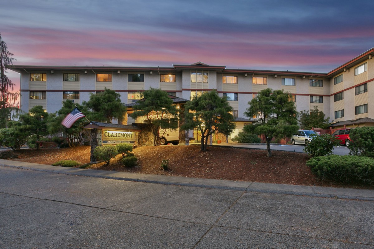 An apartment building at sunset with illuminated windows and trees in the foreground.