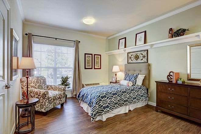 A cozy bedroom featuring a neatly made bed, a patterned armchair, and elegant decor with natural light streaming through a large window.