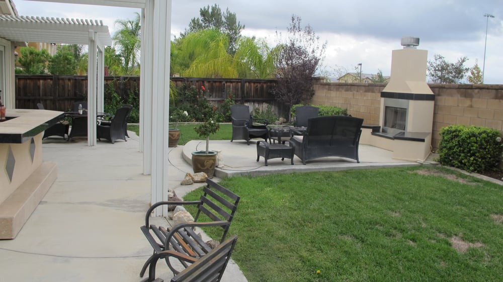 A backyard with a patio featuring outdoor seating and a fireplace, surrounded by a green lawn and garden.