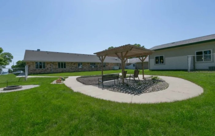 A sunny courtyard with a gazebo and outdoor seating surrounded by green grass and buildings.