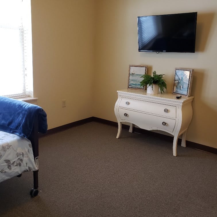 A minimalist bedroom with a mounted television, a white dresser adorned with frames and a plant, and natural light from the window.