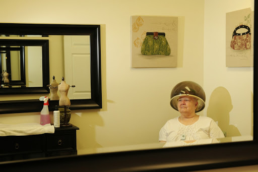 An older woman waiting under a hair dryer in a salon, reflected in the mirror.