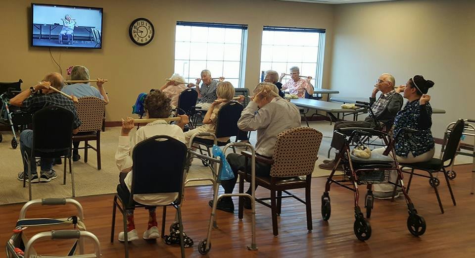 A group of elderly people gathered in a community room, some in wheelchairs, enjoying each other's company during the day.