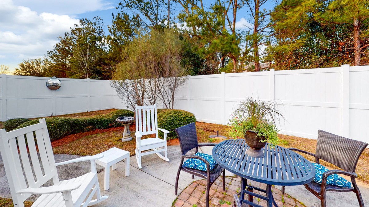 A cozy backyard patio with outdoor furniture, including a rocking chair and a table surrounded by a garden and a white fence.