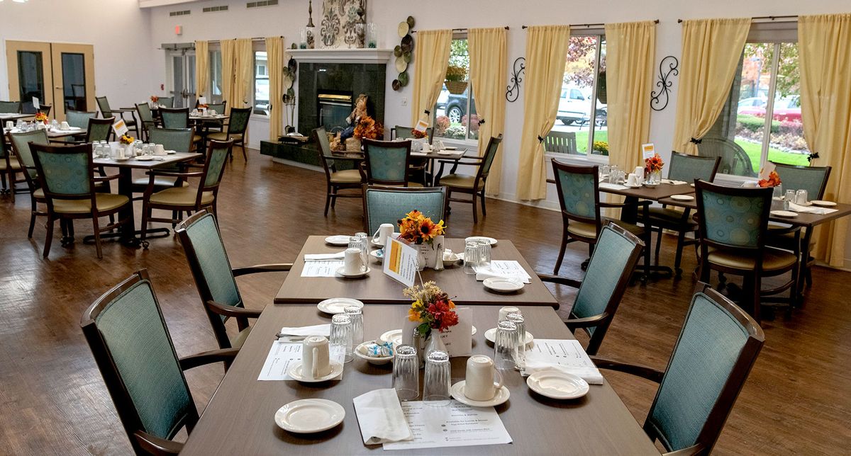 An elegantly set dining room with neatly arranged tables and chairs, accented by warm lighting and large windows with yellow curtains.