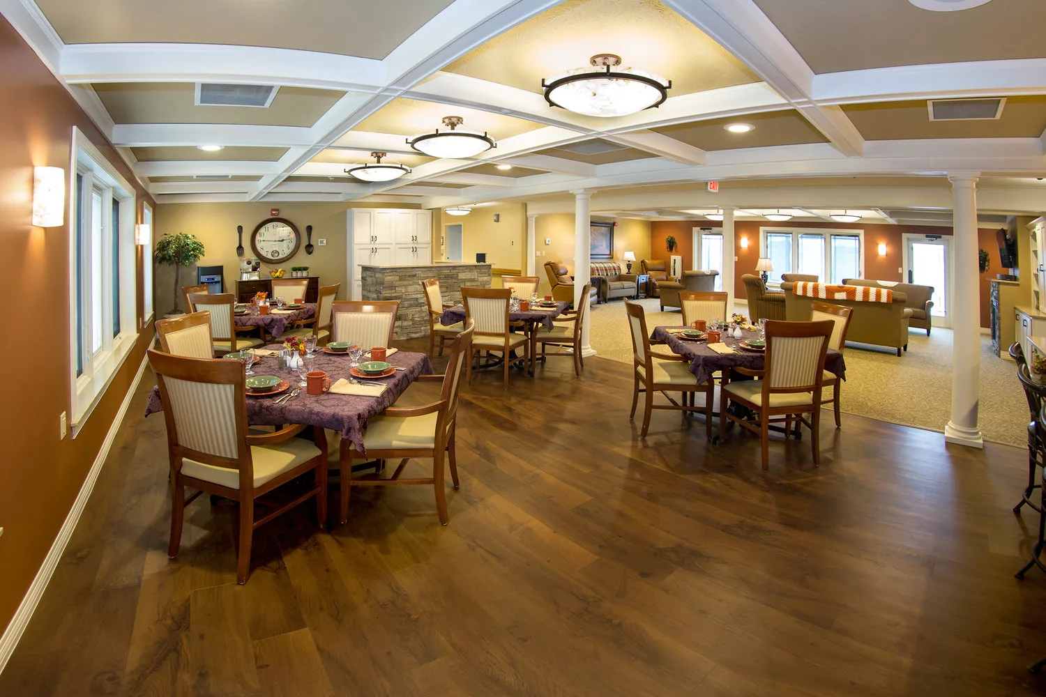 A spacious and elegantly designed dining area with wooden floors, featuring neatly arranged tables and chairs under warm lighting.