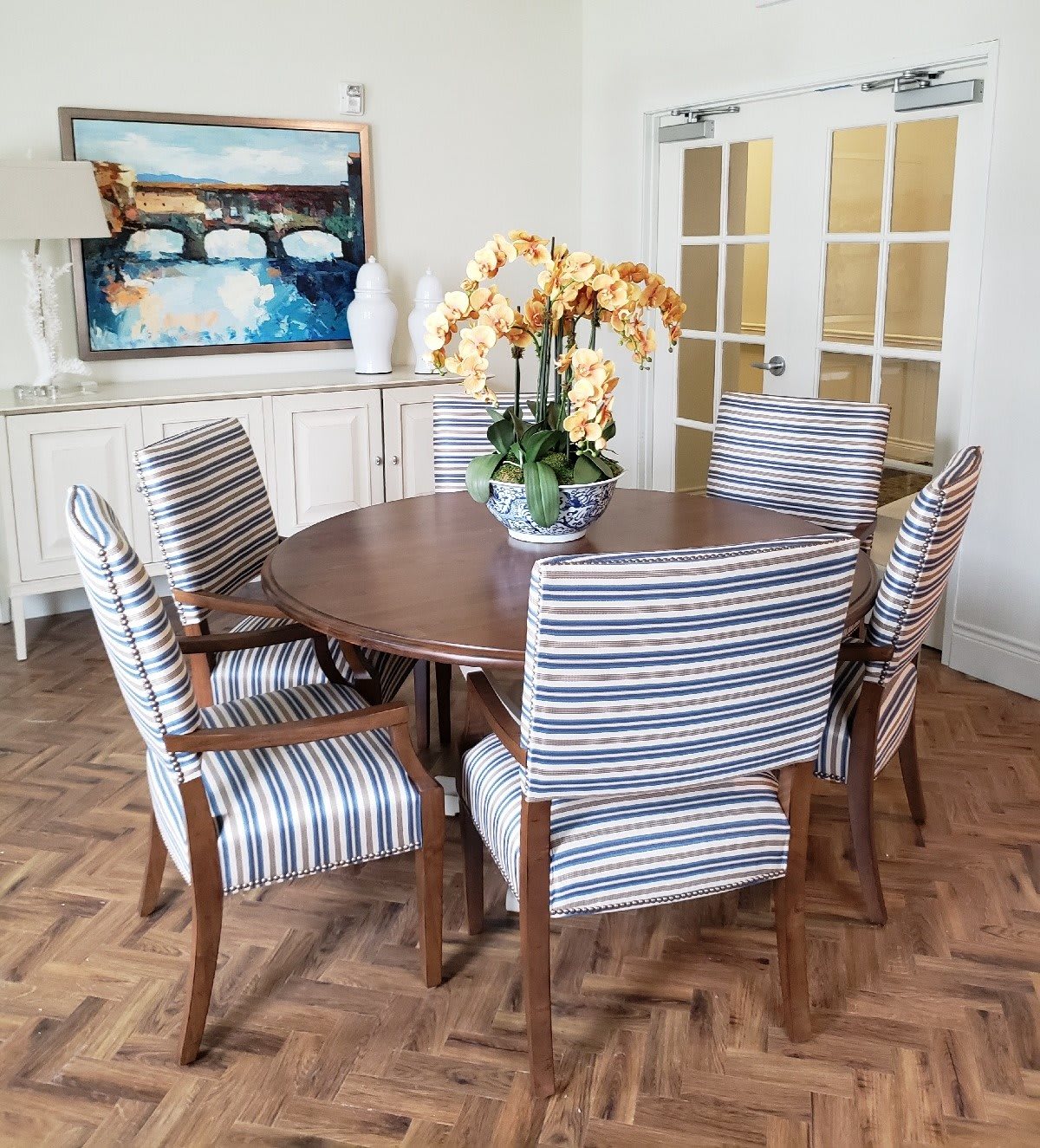 A stylish dining room featuring a wooden table surrounded by striped upholstered chairs, with a floral centerpiece and artwork on the wall.