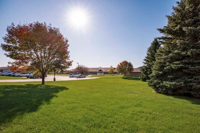 A sunny day with a grassy field, trees, and a parking lot in front of a building.