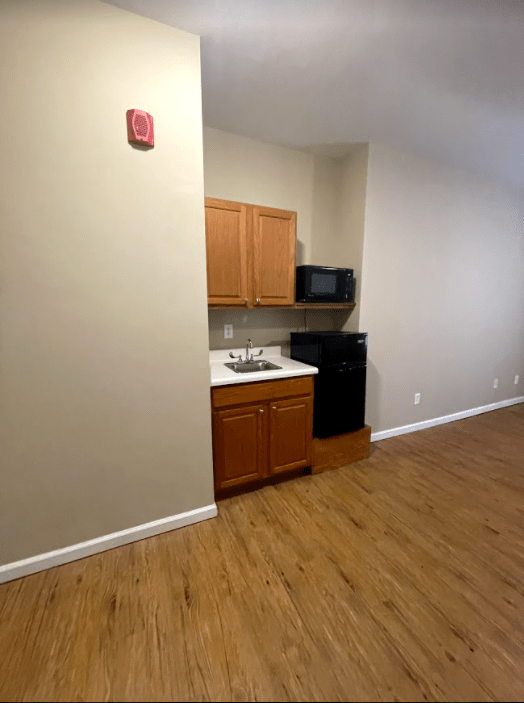 A small kitchen area in an empty apartment with wooden cabinets, a microwave, and a sink, featuring hardwood flooring.