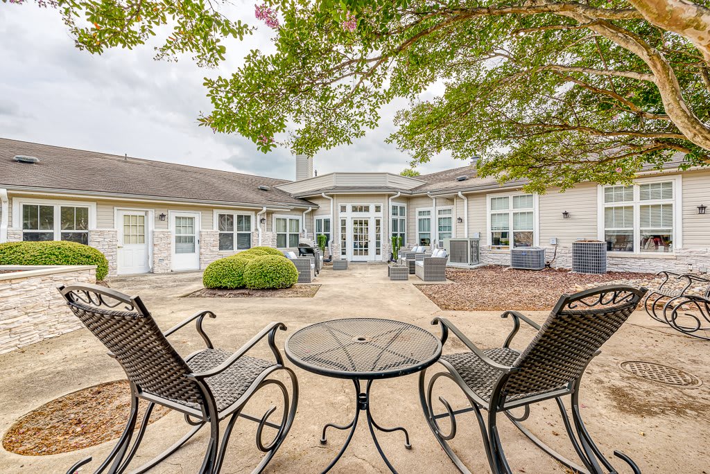 A cozy outdoor patio with metal chairs and a table, surrounded by trees and a building in the background.