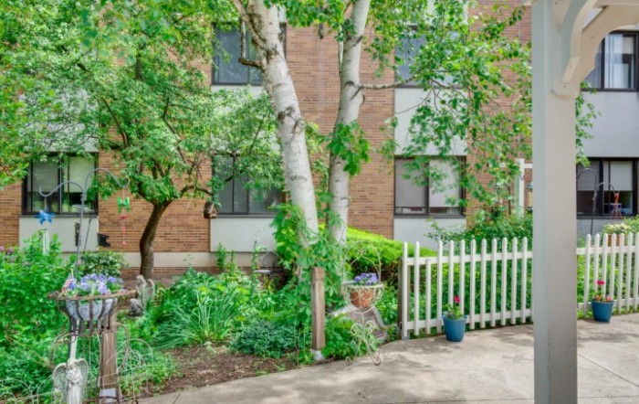 A lush garden with trees and a white picket fence in front of a brick building.