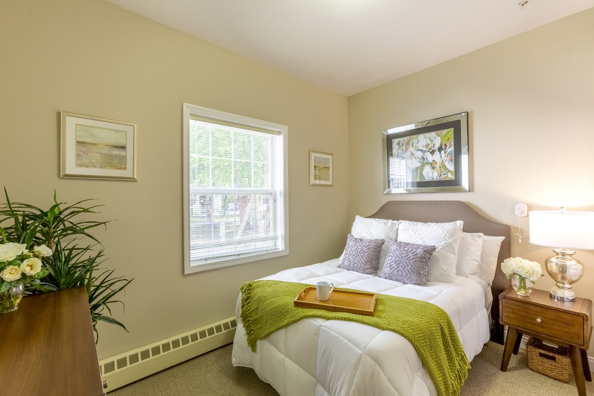 A cozy bedroom with a neatly made bed featuring green accents, framed artwork on the walls, and ample natural light from the window.