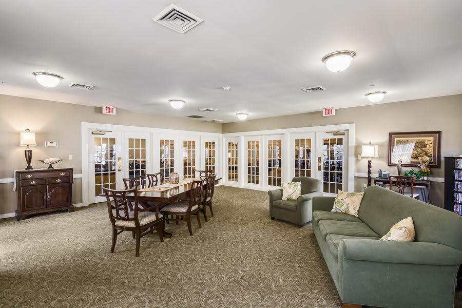 A cozy meeting room with a large dining table, chairs, and a sofa, featuring bright lighting and carpeted flooring.