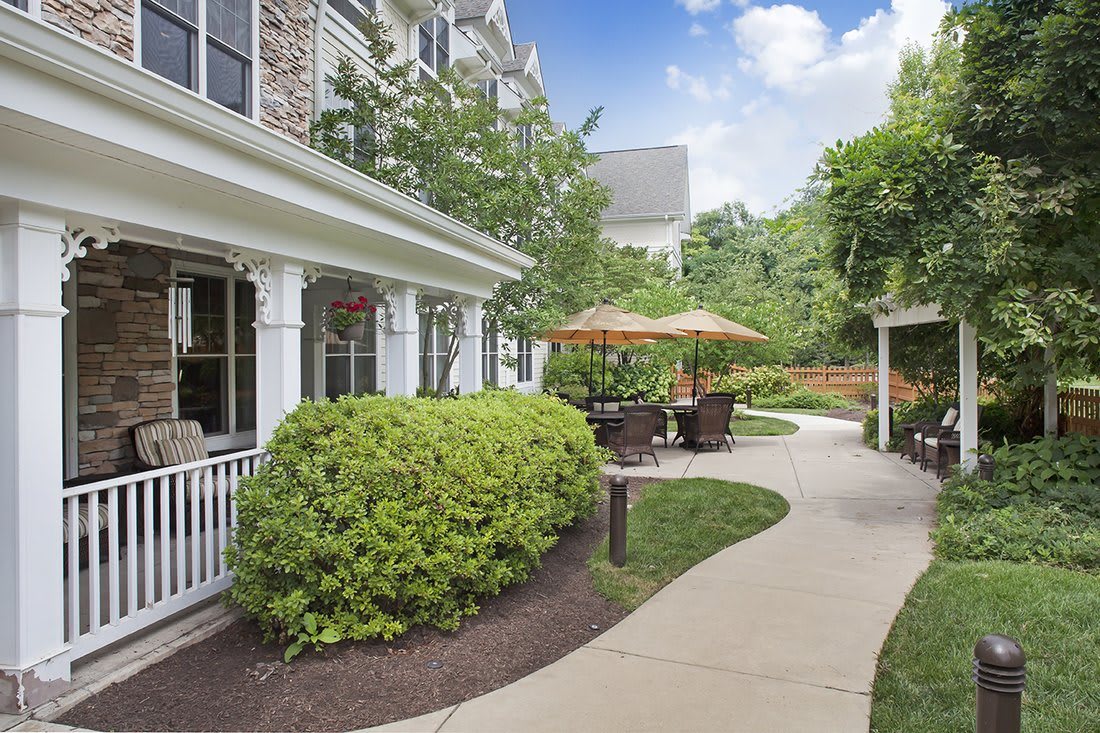 A charming garden area with a winding pathway, surrounded by greenery and featuring patio furniture under umbrellas.