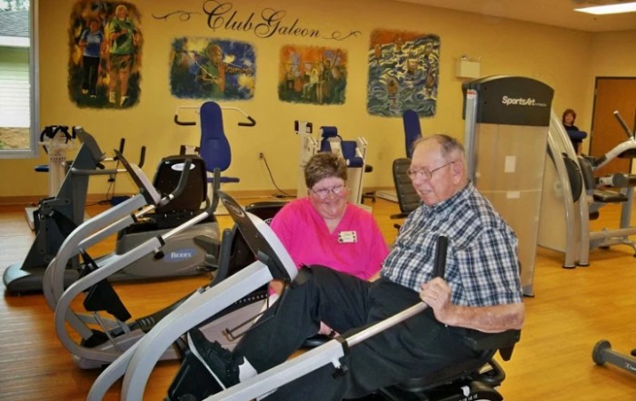 An elderly man using workout equipment with assistance from a healthcare professional in a gym setting.