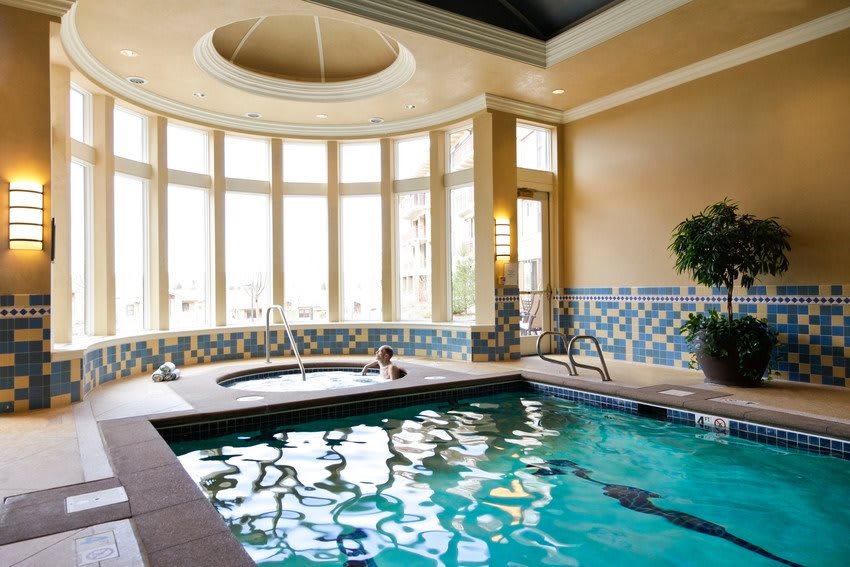 A luxurious indoor swimming pool area with a jacuzzi, surrounded by large windows allowing natural light to illuminate the space.