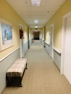 A well-lit hallway with light-colored walls and ceiling, featuring a bench and framed artwork along the sides.
