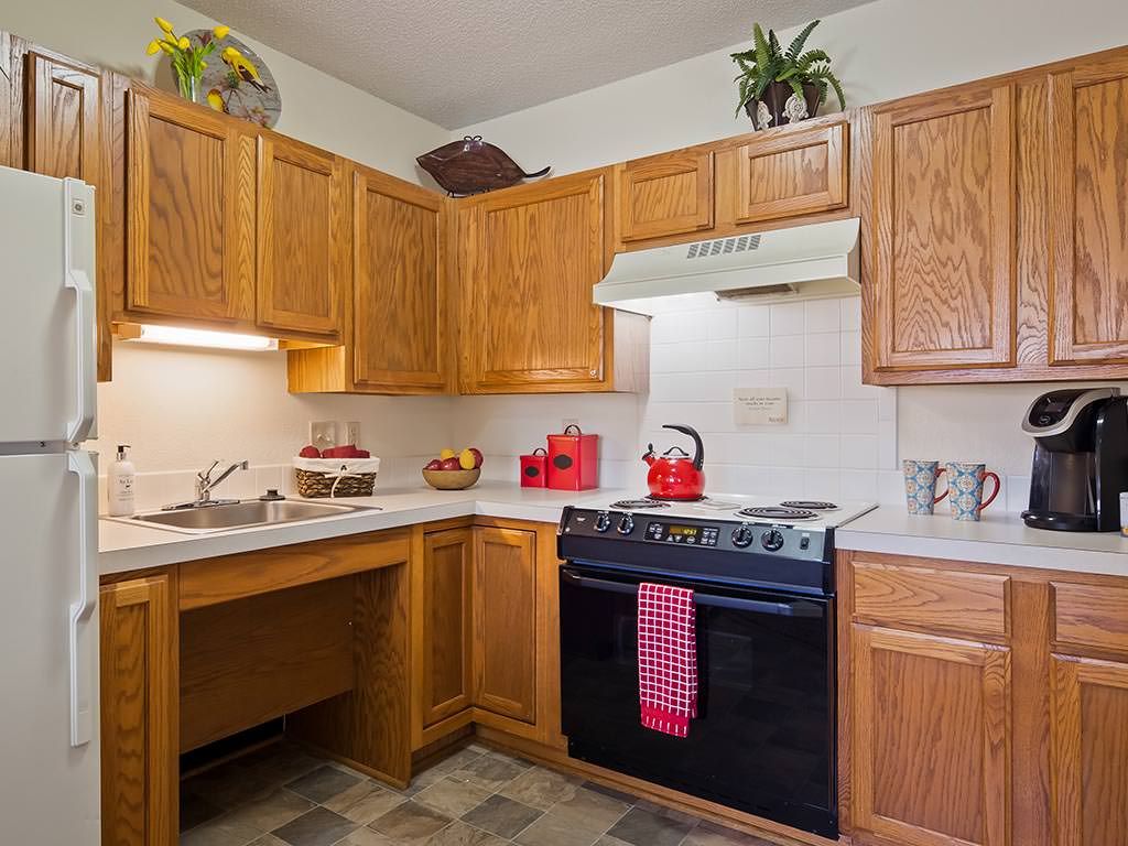 A cozy kitchen with wooden cabinets, a stove featuring a red kettle, and decorative plants.