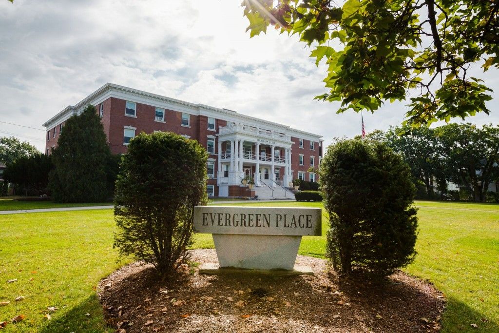 A grand brick building with white columns stands behind a sign reading 'Evergreen Place', surrounded by trees and greenery.