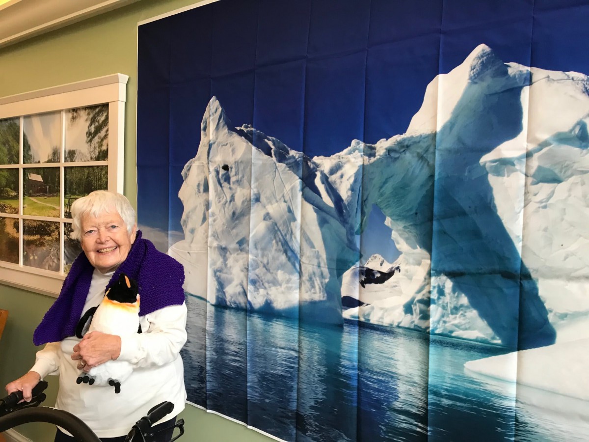An elderly person smiling in front of a large iceberg image displayed on the wall.