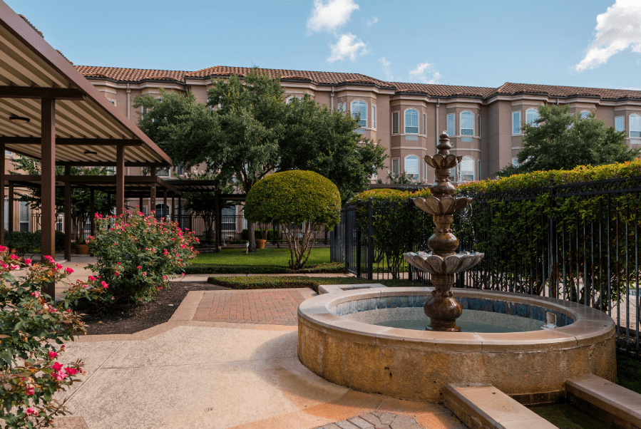 A scenic courtyard featuring a fountain surrounded by well-maintained gardens and a large building in the background.