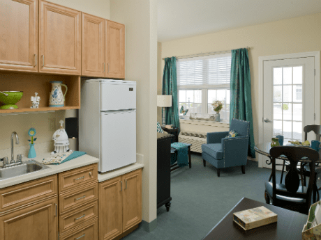 A cozy apartment interior featuring a small kitchen area and a bright living space with blue chairs and drapes.