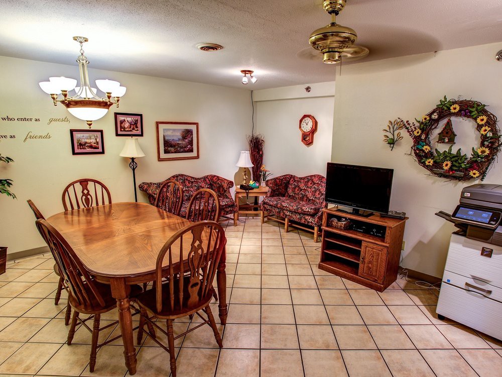 A cozy living and dining space featuring a wooden dining table with chairs, a comfortable sofa set, and a television. The room is decorated with framed pictures and has a ceiling fan for ventilation.