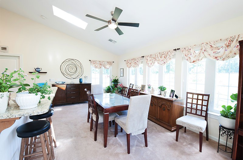 A bright dining room with a large table, surrounded by chairs and decorated with indoor plants, featuring a ceiling fan and ample natural light from the windows.