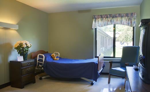 A simple hospital room with a bed, chair, and a large window allowing natural light.