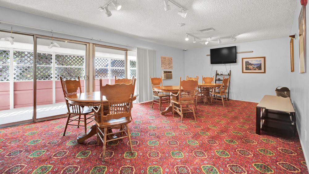 A dining room with wooden tables and chairs, featuring a patterned carpet and a wall-mounted TV.