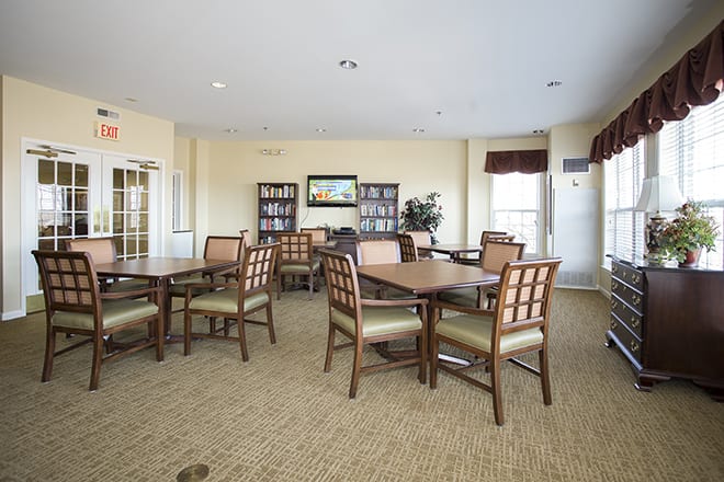 A well-lit room with multiple tables and chairs, featuring bookshelves and large windows.