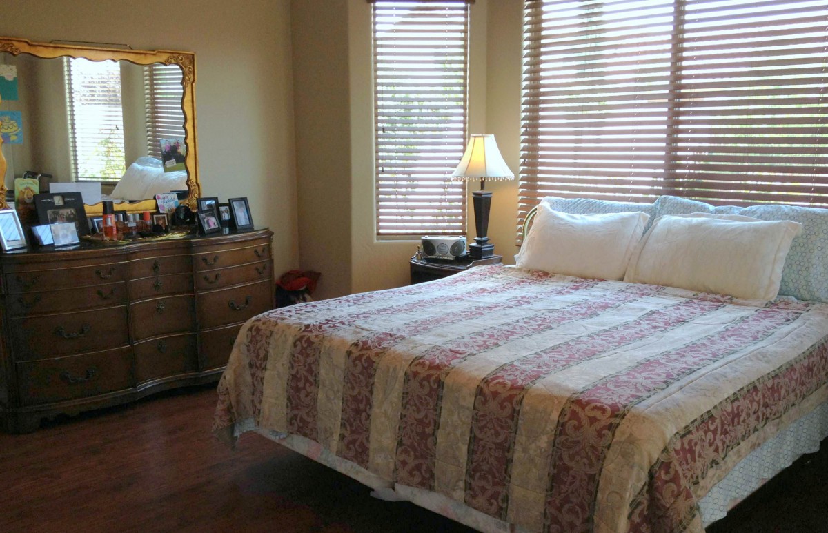 A cozy bedroom featuring a neatly made bed with a patterned quilt, next to a wooden dresser and illuminated by a table lamp near the window.