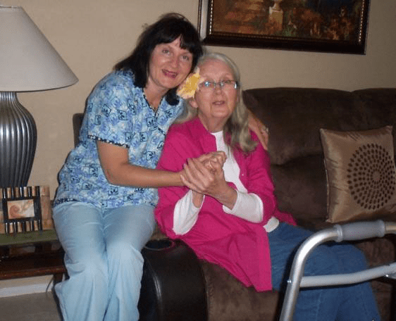 An elderly woman and a caregiver sitting together on a couch in a home setting.