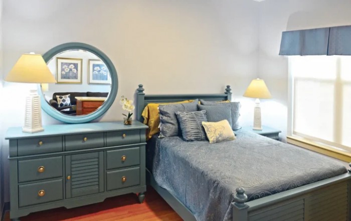 A cozy bedroom featuring a bed with blue bedding, a matching dresser with a round mirror, and a bedside lamp.