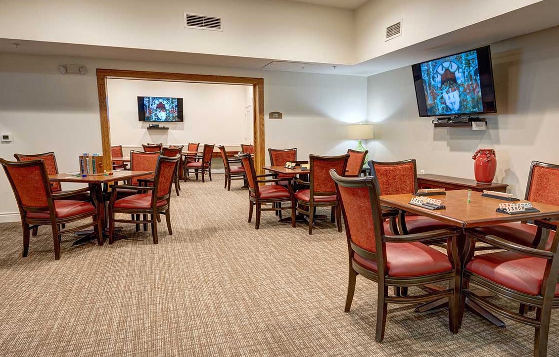 An empty restaurant dining area with wooden tables and chairs, carpeted flooring, and TV screens mounted on the walls.