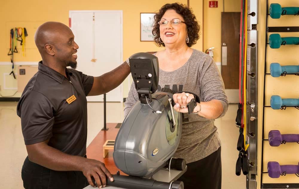 A woman uses an exercise bike while a trainer assists her in the gym.