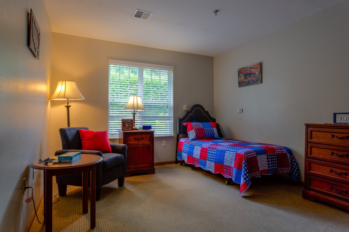 A cozy bedroom featuring a twin bed with a colorful quilt, a comfortable chair, and a warm lamp by the window.