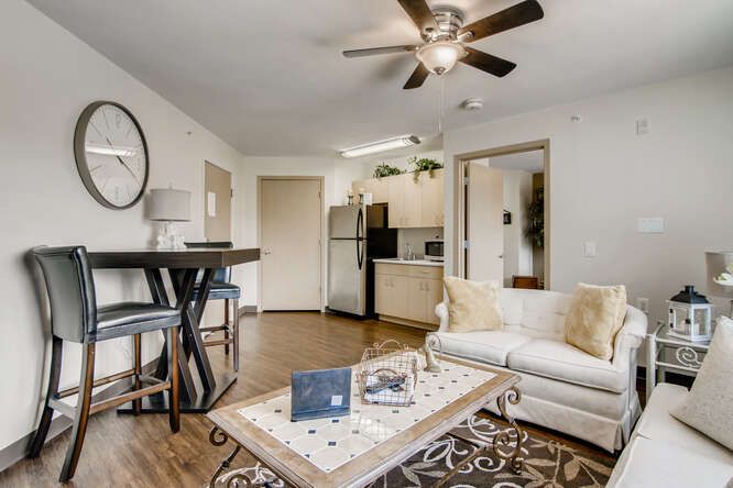 A modern, open-concept living room and kitchen area featuring a ceiling fan, sofa, bar stools, and hardwood flooring.