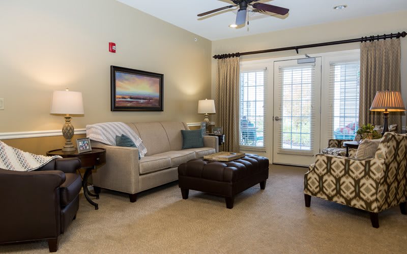 A cozy living room with a sofa, armchair, and a coffee table under a ceiling fan, featuring artwork on the wall and natural light from french doors.