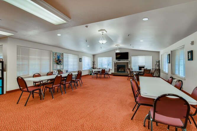 A spacious meeting room with tables and chairs arranged on an orange carpet, featuring large windows and ample lighting.