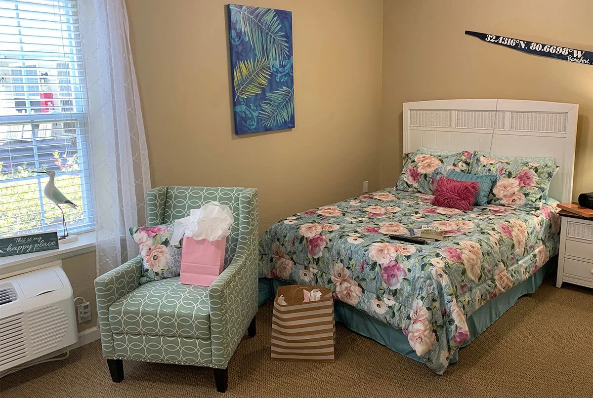 A cozy bedroom featuring a bed with floral bedding and a matching armchair, decorated with a colorful painting on the wall.