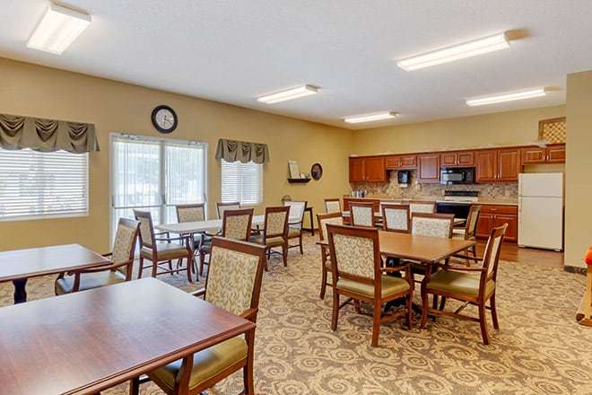 A well-lit dining room with tables and chairs, featuring a kitchen area in the background.