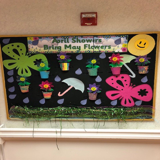 A spring-themed bulletin board with the phrase 'April Showers Bring May Flowers,' featuring colorful flowers, butterflies, and an umbrella.