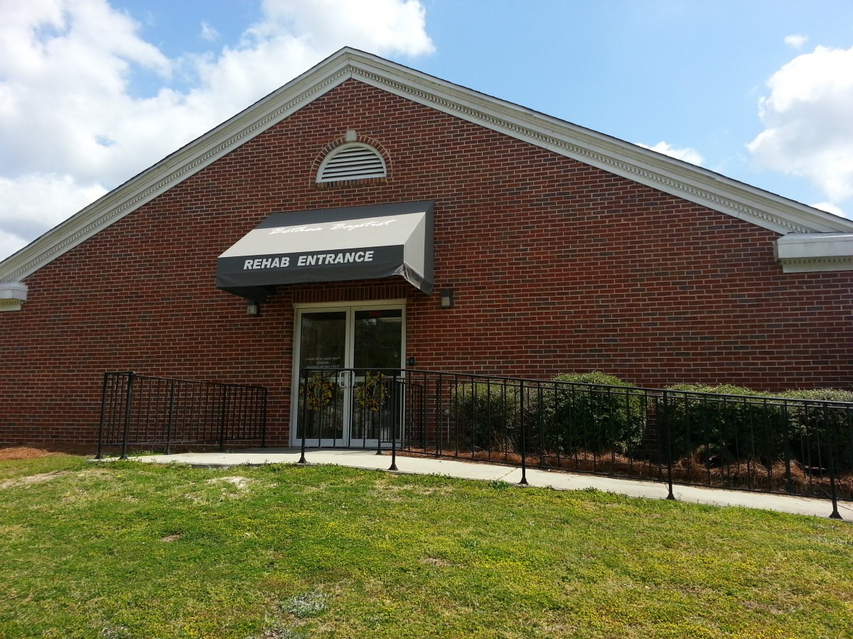 A brick building with an awning over the entrance, surrounded by grass and shrubs.