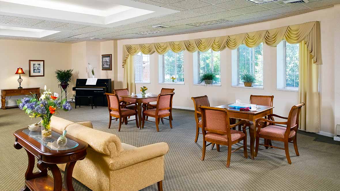 A cozy and spacious living area with multiple tables, chairs, a sofa, and a piano by large windows adorned with curtains.
