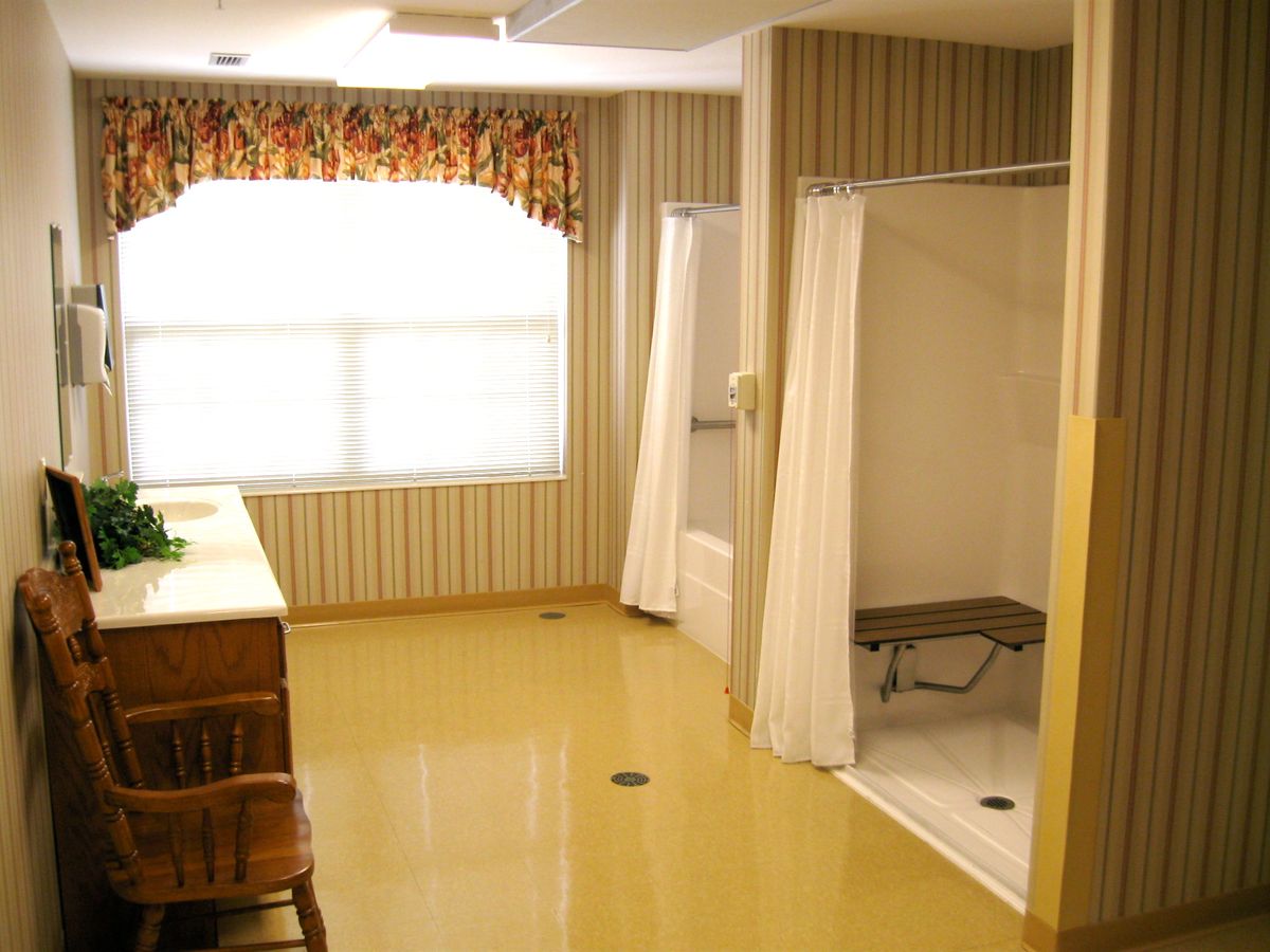 A spacious bathroom with a shower area, wooden chair, sink, and window with floral curtains.