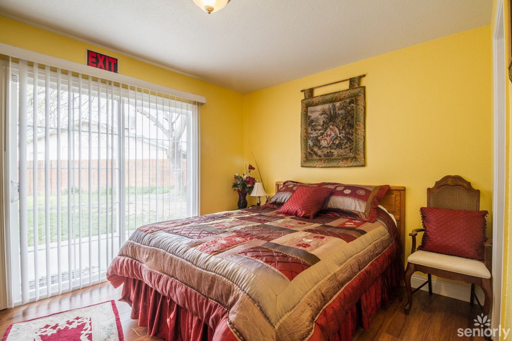 A cozy bedroom with yellow walls featuring a quilted bedspread and decorative pillows, illuminated by natural light coming through a sliding glass door.