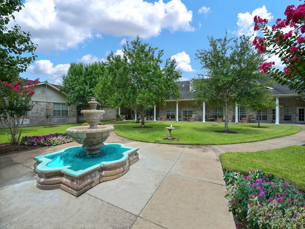A beautiful garden featuring a central fountain surrounded by trees and flowers under a bright blue sky.