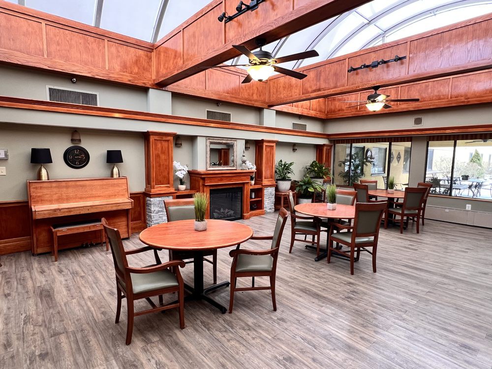 A spacious, well-lit interior with wooden furniture, featuring tables, chairs, a piano, a fireplace, and decorative plants.